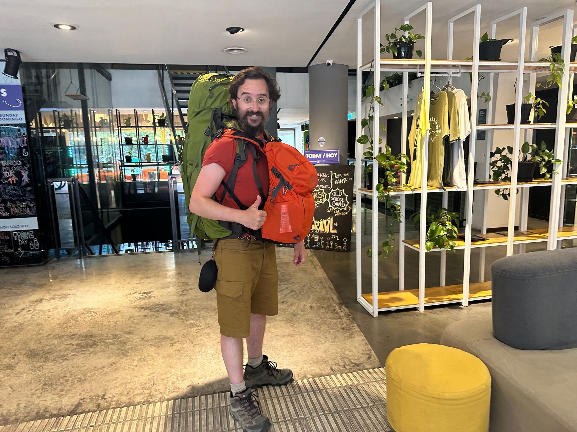 A photo of JP with both backpacks on, giving a thumbs up, in a hostel lobby.